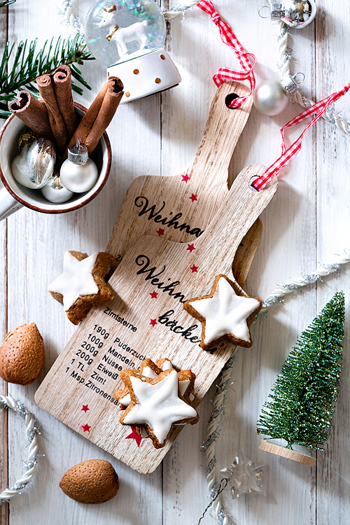 Zimtsterne Weihnachtsplätzchen liegen auf kleinen Holzbrettchen, die mit dem Rezept der Zimtsterne bedruckt sind. Ringsherum liegt schöne Weihnachtsdeko wie ein grüner Tannenbaum aus Draht, eine Espresso-Tasse mit Weihnachtskugeln und eine Schneekugel mit Hirsch, sowie ein paar Mandeln.
