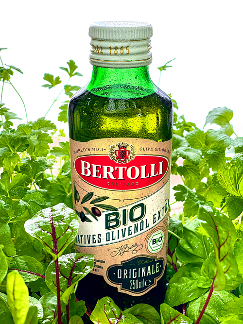 Eine Flasche Bertolli natives bio Olivenöl extra mit Wassertropfen steht inmitten von frischen Kräutern wie Petersilie, Basilikum, Zitronenmelisse und Sauerampfer im Gegenlicht vor weißem Hintergrund in der Küche.