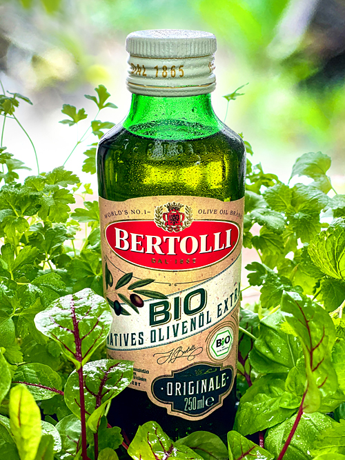 Eine Flasche Bertolli natives bio Olivenöl extra mit Wassertropfen steht inmitten von frischen Kräutern wie Petersilie, Basilikum, Zitronenmelisse und Sauerampfer im Gegenlicht vor grünem Gartenhintergrund