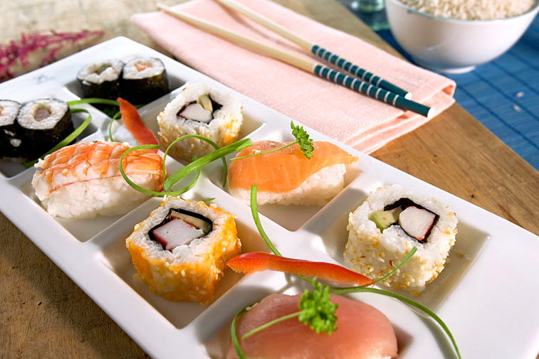 Auf dem Bild ist eine vielfältige Auswahl an Sushi zu sehen: Nigiri-Sushi mit Lachs oder Garnelen, sowie Ura-Maki-Rollen mit Surimi, Avocado oder Gurke. Die Maki-Sushi-Röllchen, umhüllt von Nori-Algenblättern, sind auf einem Porzellanteller auf einem Holzbrett arrangiert. Neben dem Teller liegen eine lachsfarbene Stoffserviette und Essstäbchen mit blau lackierten Griffen. Im Hintergrund steht eine Schale mit Reis.